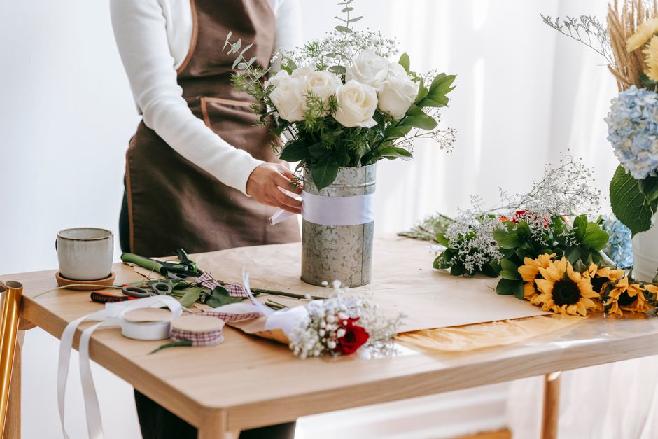 A person holding a bouquet of white roses and greenery, assembled in a textured silver vase wrapped with a white ribbon, is positioned at a wooden table. The workspace displays various floral arrangin